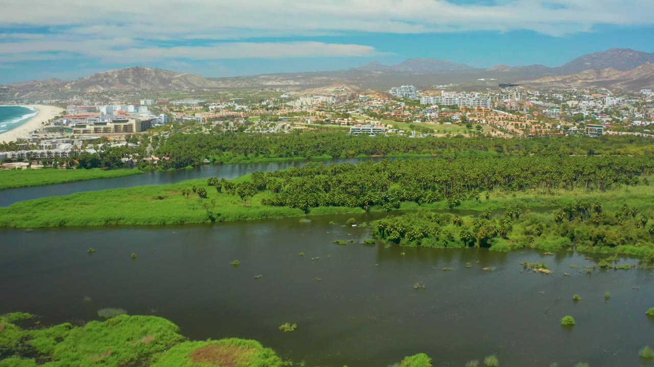 Aerial frame over San Jose Del Cabo showing broad river water below, dense green vegetation across the midground, city buildings and beach in the distance, mountains on the horizon, and no visible people.