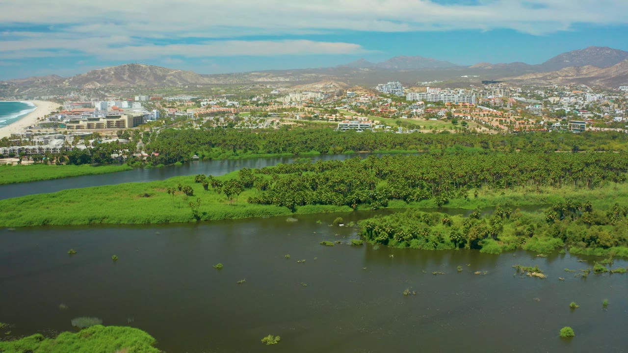 Wide San Jose Del Cabo aerial showing river water across the foreground, a long green vegetated band, city blocks and beach beyond, mountain ridges under blue sky, and no visible people.