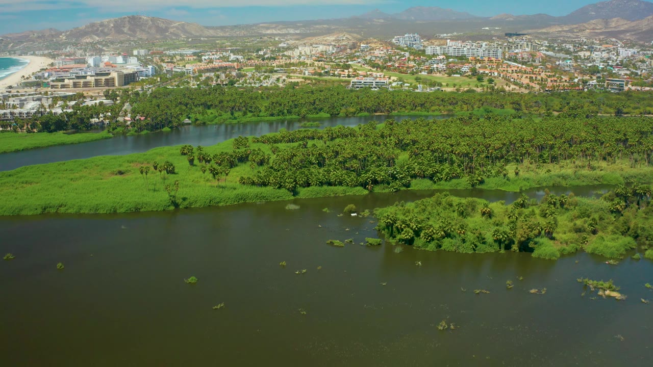 Wide San Jose del Cabo frame showing dark green river water at bottom, bright vegetation and islands through the center, built-up city areas and a beach edge in the distance, mountains behind, and no visible people.