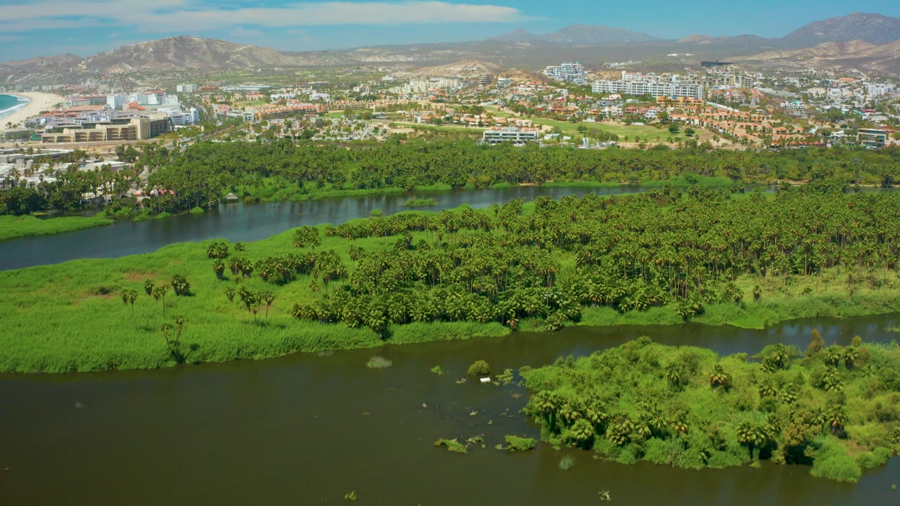 Aerial frame over San Jose del Cabo showing dark river water in the foreground, dense green vegetation across the center, city buildings beyond, mountains on the horizon, and no visible people.