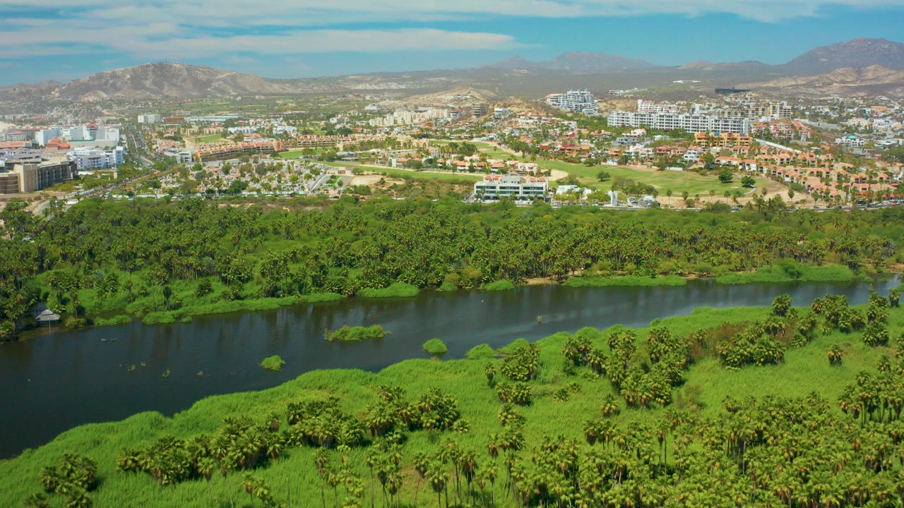 Opening aerial frame over San Jose del Cabo shows a dark river bordered by dense green vegetation, urban buildings beyond it, distant mountains, blue sky, and no visible people.