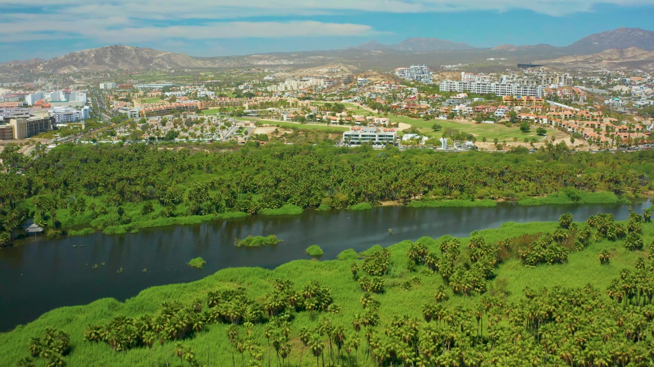 At one second, the San Jose del Cabo aerial view holds on the river and bright green banks, with white buildings and brown mountains under a lightly clouded sky, no visible people.
