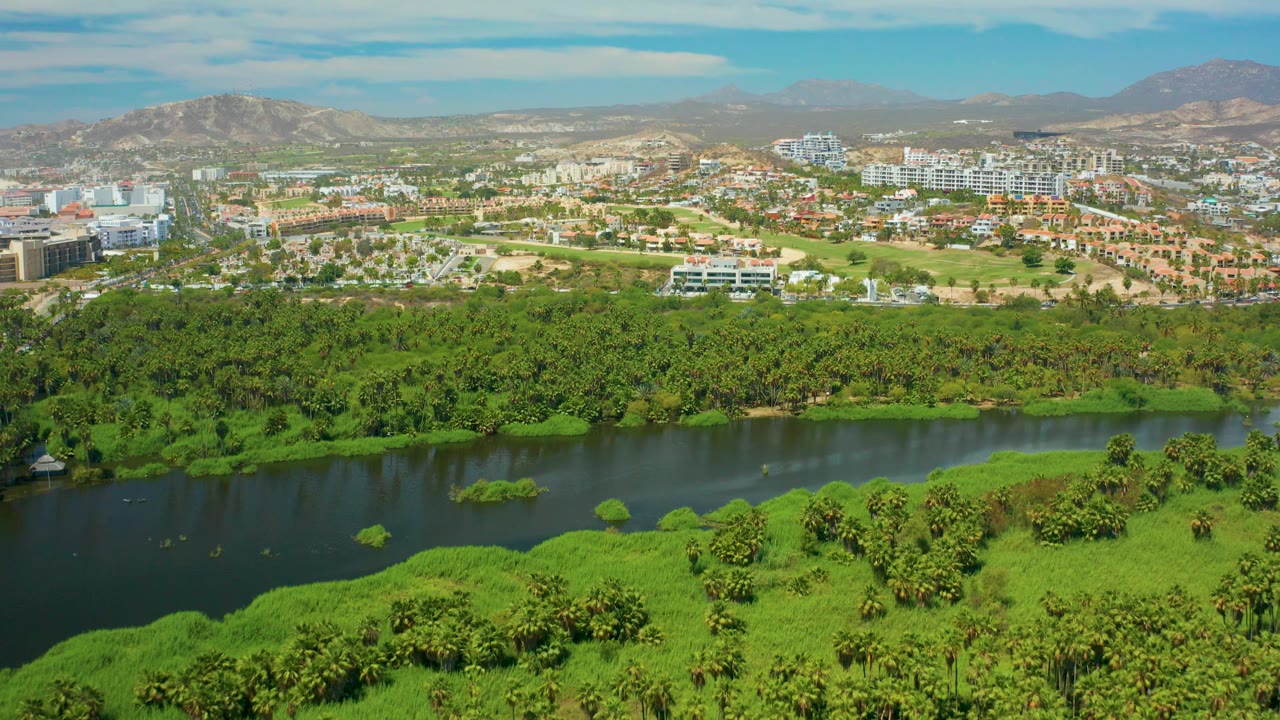 At two seconds, the frame shows the San Jose del Cabo river corridor across the lower half, compact urban development in the distance, hazy mountains, scattered clouds, and no visible people.