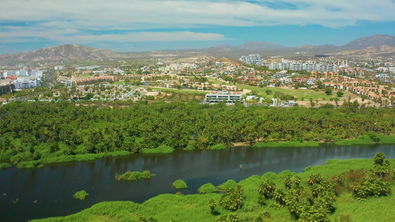 Wide 0:00 view over San Jose del Cabo with dark river water in the foreground, dense green vegetation, distant town buildings, tan mountains, blue sky, and no visible people.