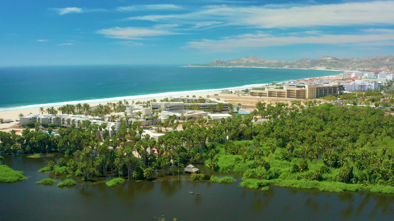Aerial view in San Jose Del Cabo showing dark lagoon water with green floating vegetation in front of a sandy beach, blue ocean, beachfront buildings, distant hills, and no visible people.