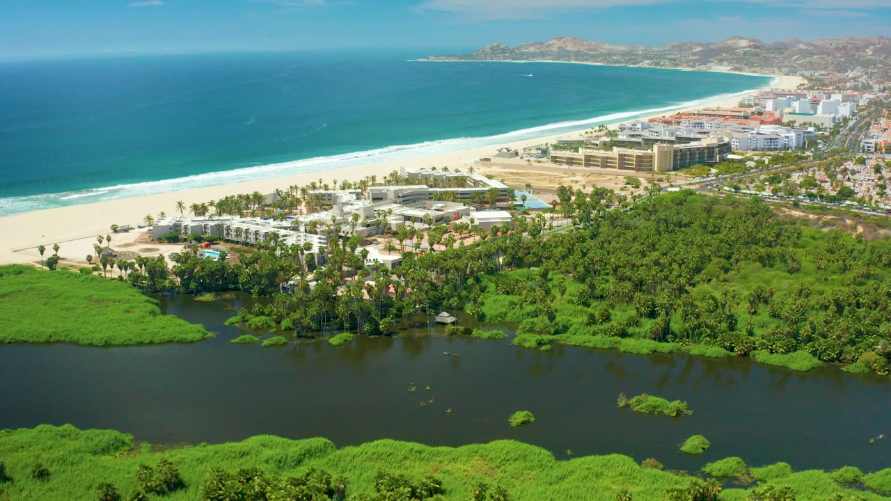 Opening aerial frame over San Jose Del Cabo shows a dark lagoon in the foreground, dense green vegetation, a sandy beach, blue ocean, distant hills, and no visible people.