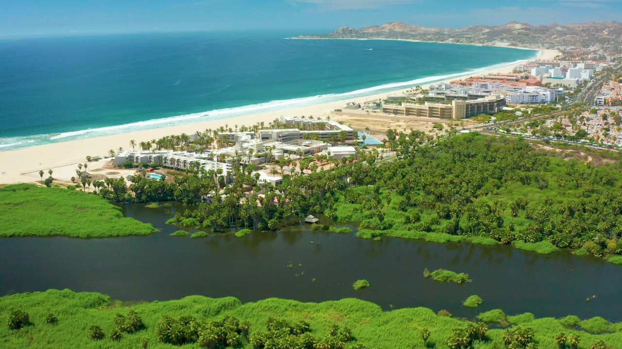 At 0:03, San Jose Del Cabo beachside buildings and palms sit between the sandy shore and green lagoon vegetation, with blue water beyond and no visible people.
