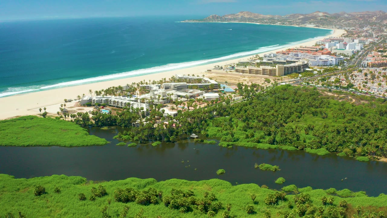 Opening aerial frame over San Jose del Cabo shows dark lagoon water edged by green vegetation, a pale beach, resort buildings, blue ocean, white surf, distant hills, and no visible people.