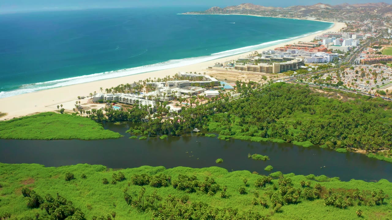 At 0:01, the San Jose Del Cabo coastline remains in a wide aerial composition, with the lagoon band below, bright beach and surf across the middle, buildings and roads near shore, and no visible people.