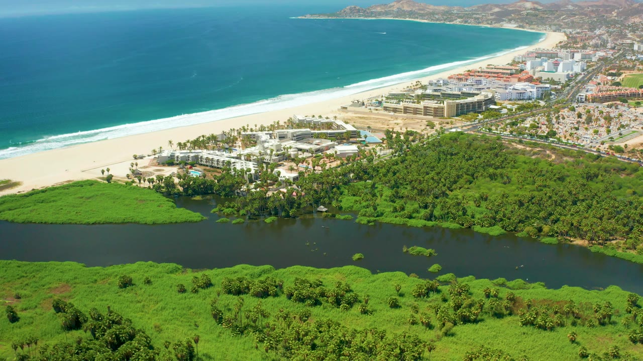 At 0:03, the aerial view over San Jose Del Cabo shows the dark lagoon stretching across the lower frame, thick green brush, a pale sandy beach, rolling ocean waves, nearby buildings, and no visible people.