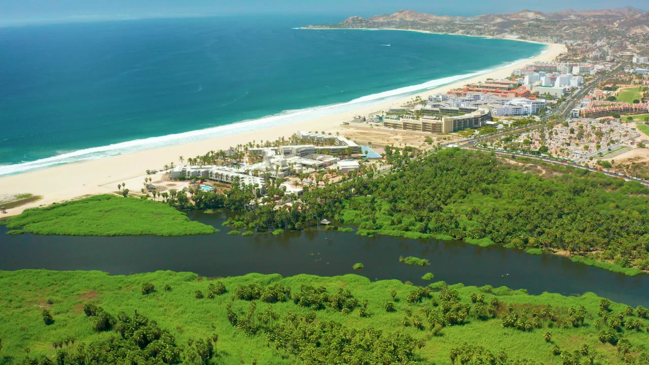 High aerial view of San Jose Del Cabo with turquoise ocean at left, white surf along the beach, a dark lagoon and green vegetation in the foreground, beachfront buildings beyond, and no visible people.