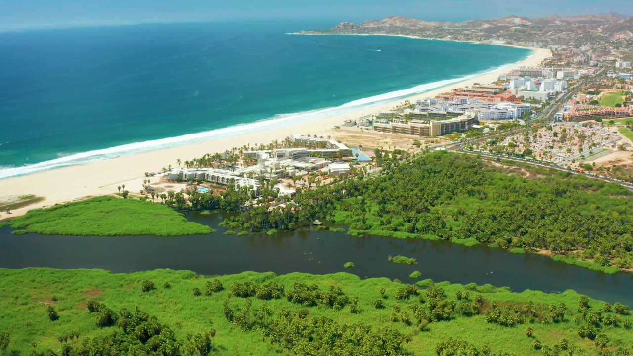Aerial view over San Jose Del Cabo showing the ocean and long surf line across the upper frame, the lagoon and bright green vegetation below, hotel-like beachfront blocks to the right, and no visible people.