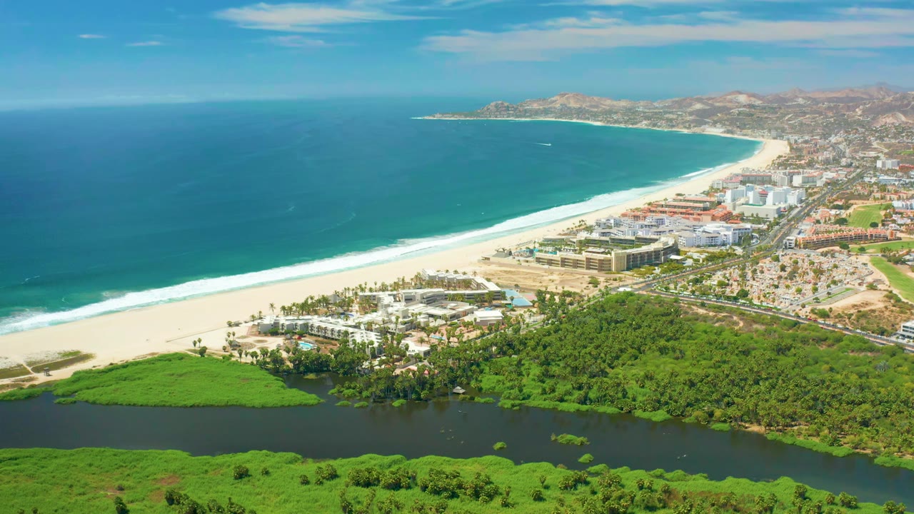 Wide San Jose Del Cabo aerial frame shows the coastline curving past a sandy beach, with ocean on the left, buildings on the shore, river water below, and no visible people.