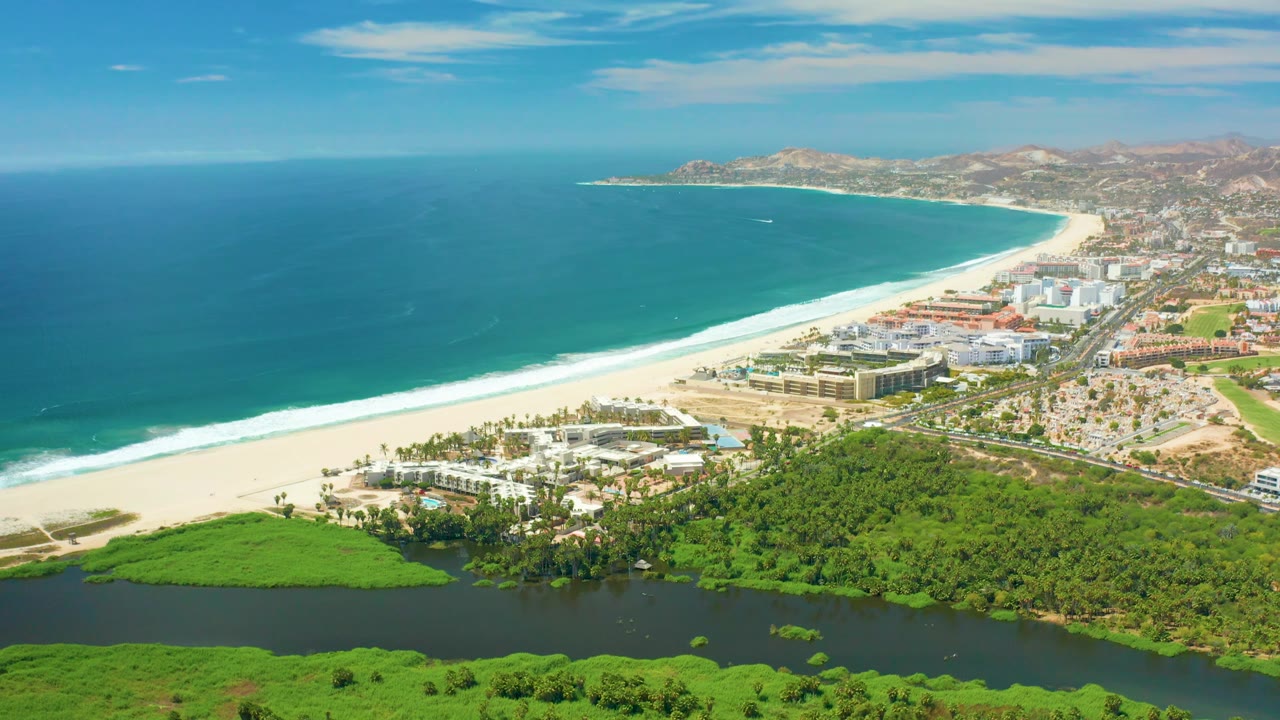 San Jose Del Cabo coastline appears from above with a long pale beach, blue-green ocean, compact shoreline development, foreground river and vegetation, distant hills, and no visible people.