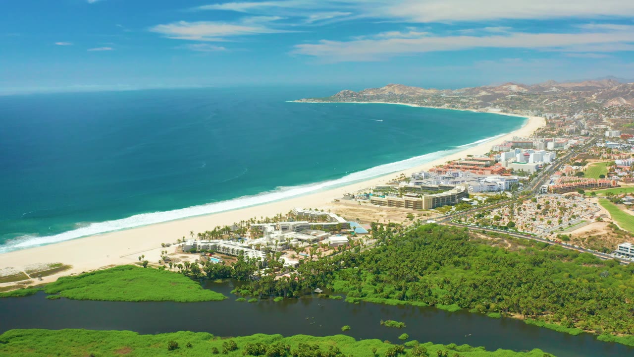 Aerial view in San Jose Del Cabo shows resort-scale buildings near the beach, a broad band of surf, dark river water and greenery in front, hills in the distance, and no visible people.