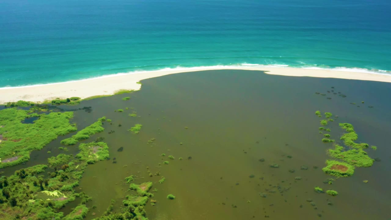 Second aerial frame in San Jose del Cabo showing the lagoon surface with small mangrove patches, a narrow white beach, blue ocean, and no visible people.