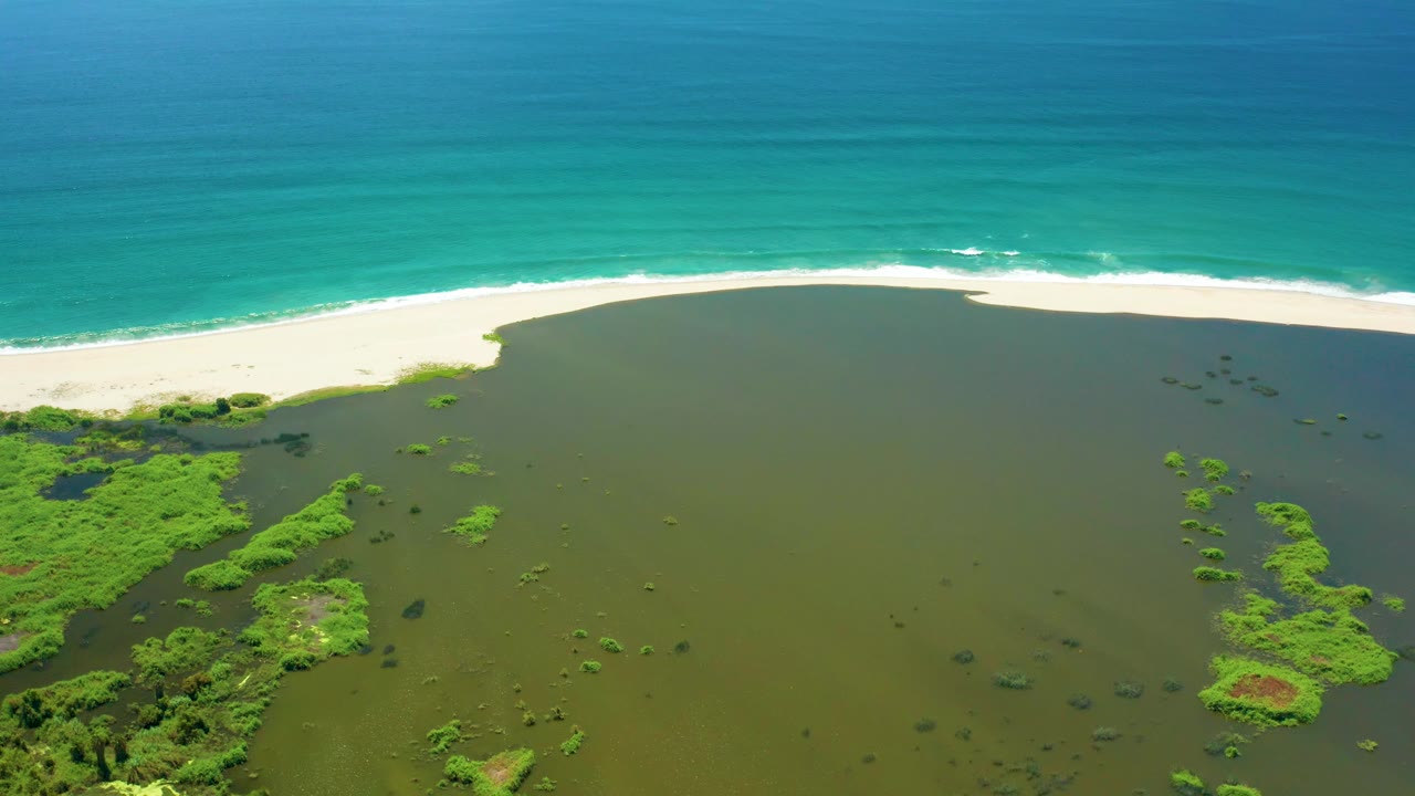 San Jose del Cabo aerial frame with the lagoon occupying most of the foreground, scattered green vegetation patches, a white beach line, and calm turquoise ocean beyond, with no visible people.
