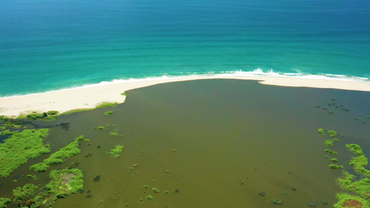 Aerial view in San Jose Del Cabo with the lagoon occupying the lower frame, bright green vegetation clustered at left, white sand through the center, ocean surf above, and no visible people.