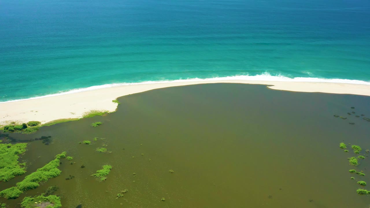 Aerial San Jose del Cabo frame with the lagoon occupying the lower half, mangrove vegetation at the left edge, a white sand strip, and turquoise ocean beyond, with no visible people.