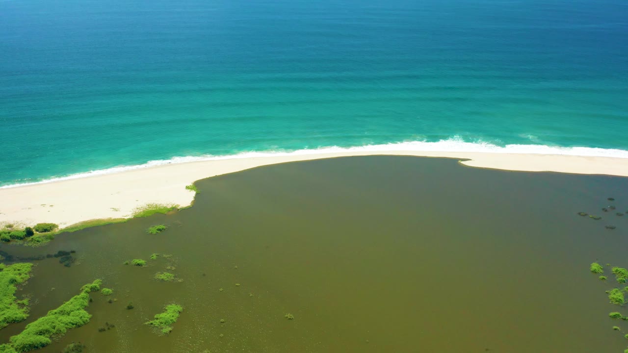 Aerial frame over San Jose del Cabo with broad green lagoon water in front, small mangrove clusters near the lower left, a sandy beach, and bright turquoise ocean, with no visible people.