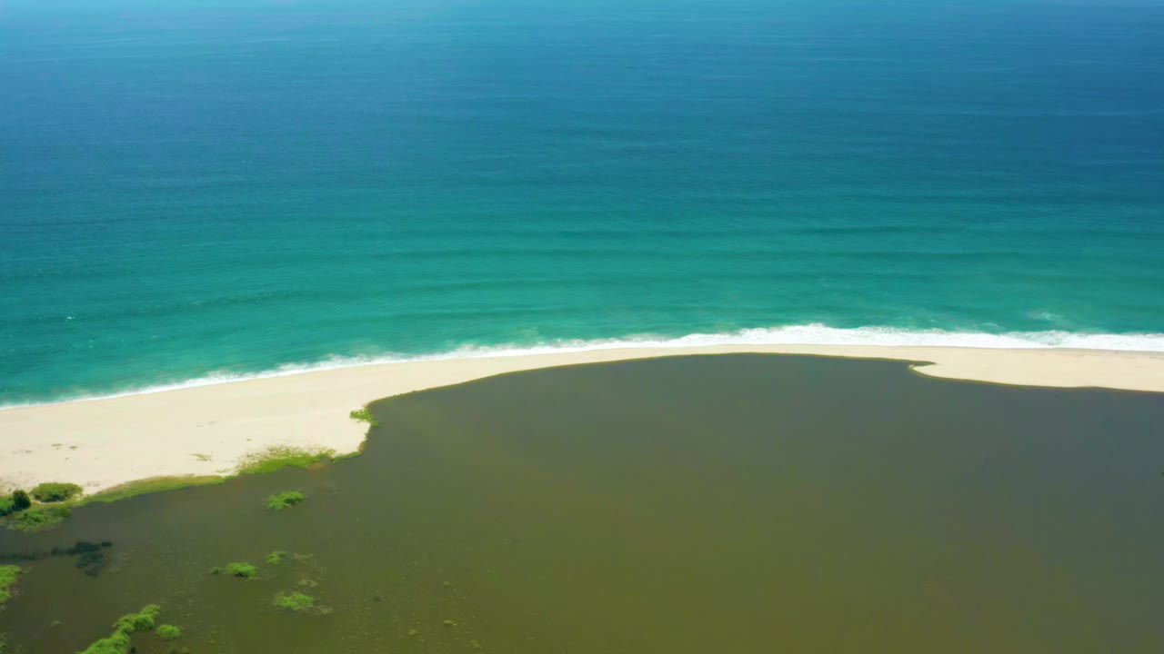 Aerial view in San Jose del Cabo with the lagoon filling the lower frame, a curved sand beach across the middle, ocean waves at the top, and no visible people.