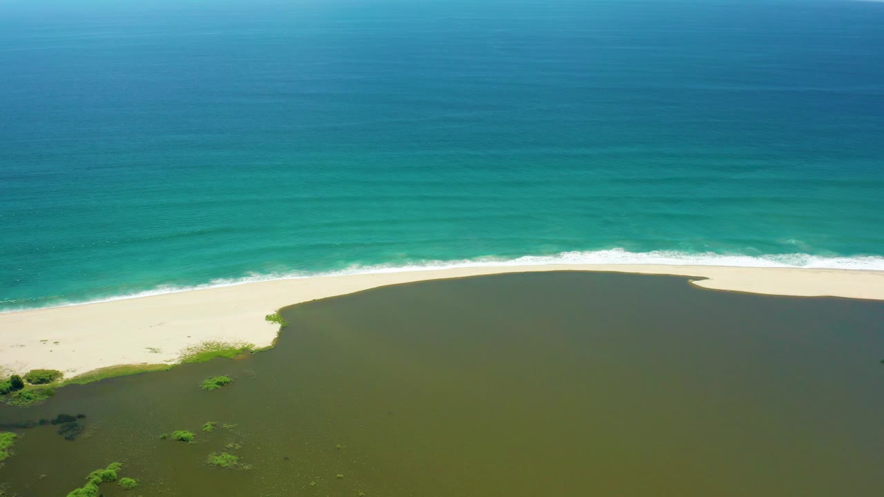 Aerial shot over San Jose del Cabo coast with green lagoon water in the foreground, a light sand barrier, turquoise ocean in the background, and no visible people.