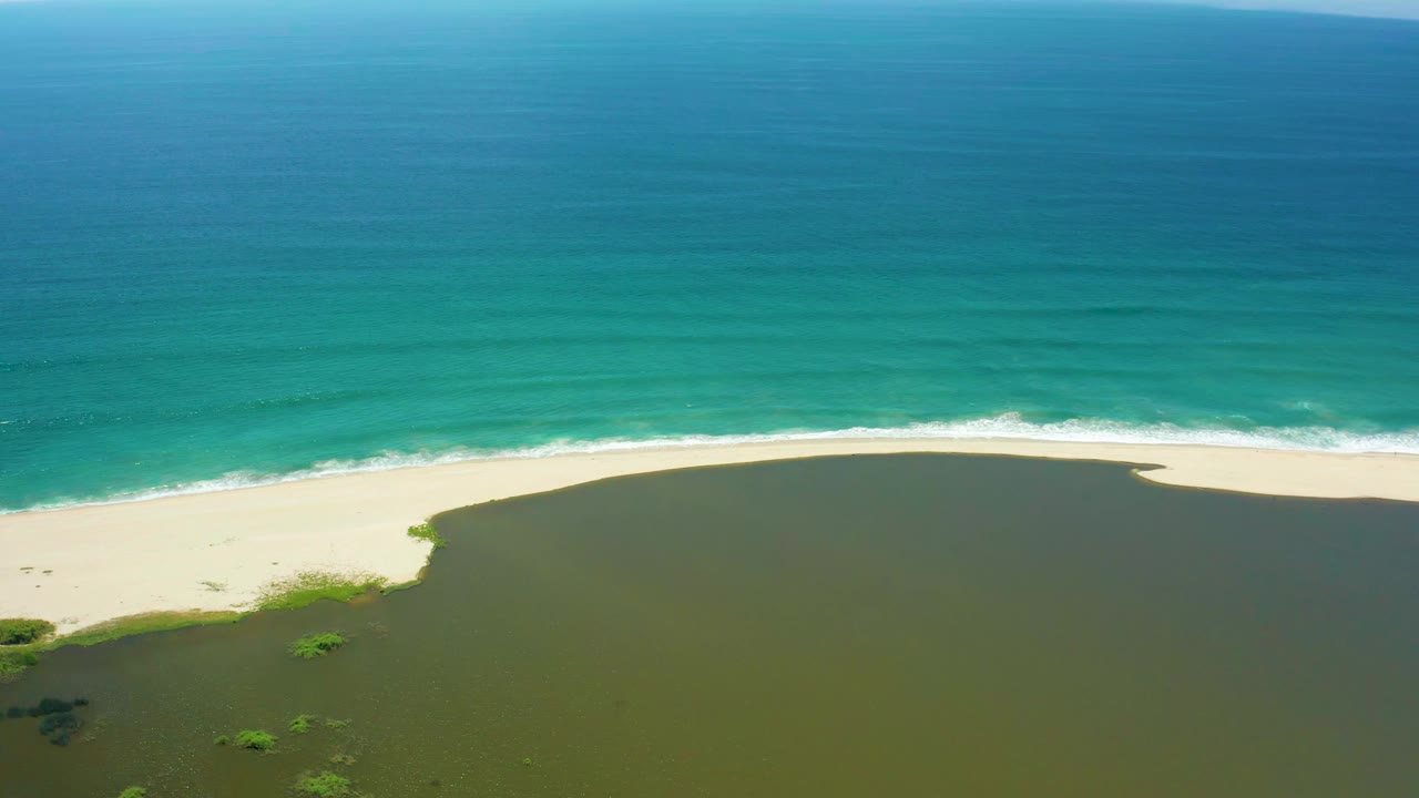 Aerial view in San Jose Del Cabo with a broad turquoise sea, a thin white surf line, pale sand, and dark green lagoon water below, with no visible people.