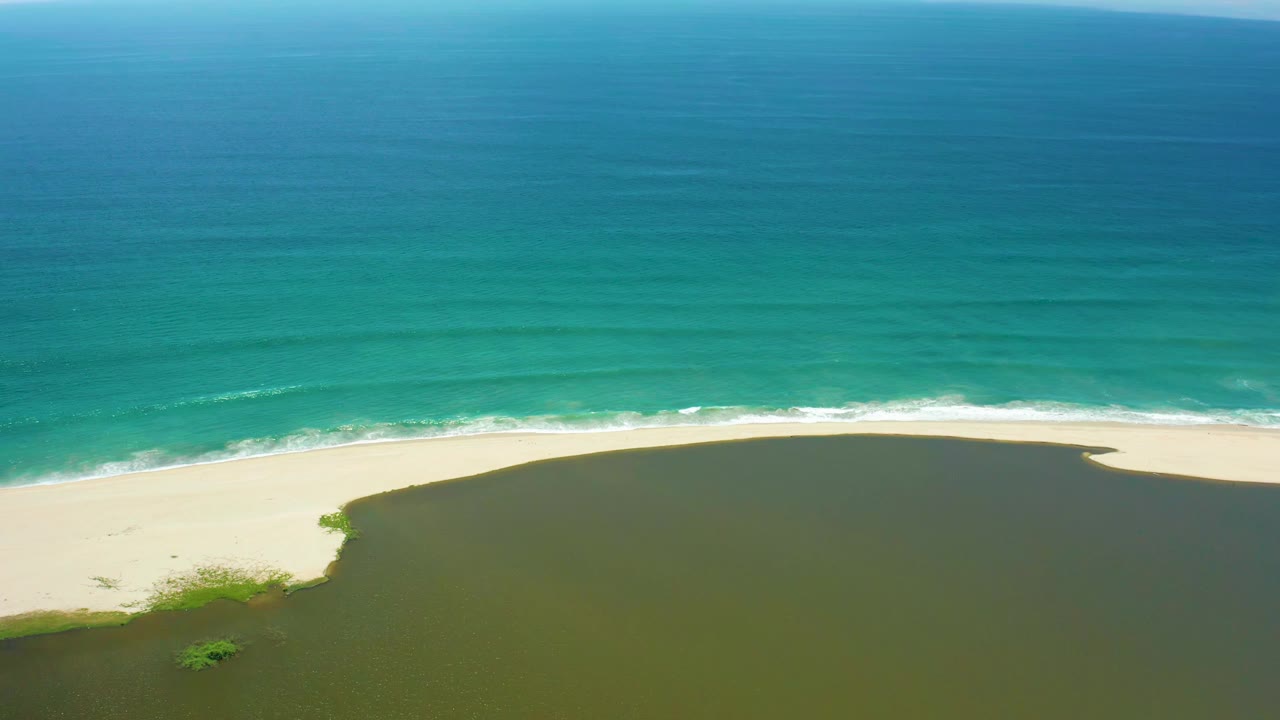 Aerial frame in San Jose Del Cabo with the ocean occupying the upper half, a thin breaking surf line along the sand, and green river water curving across the lower frame, with no visible people.