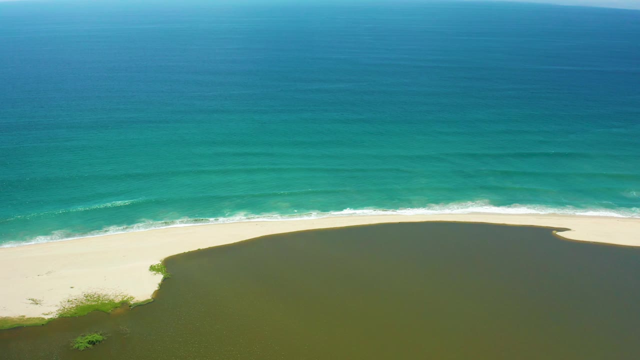 High aerial view of blue-green ocean, white shore break, sand, and river water.