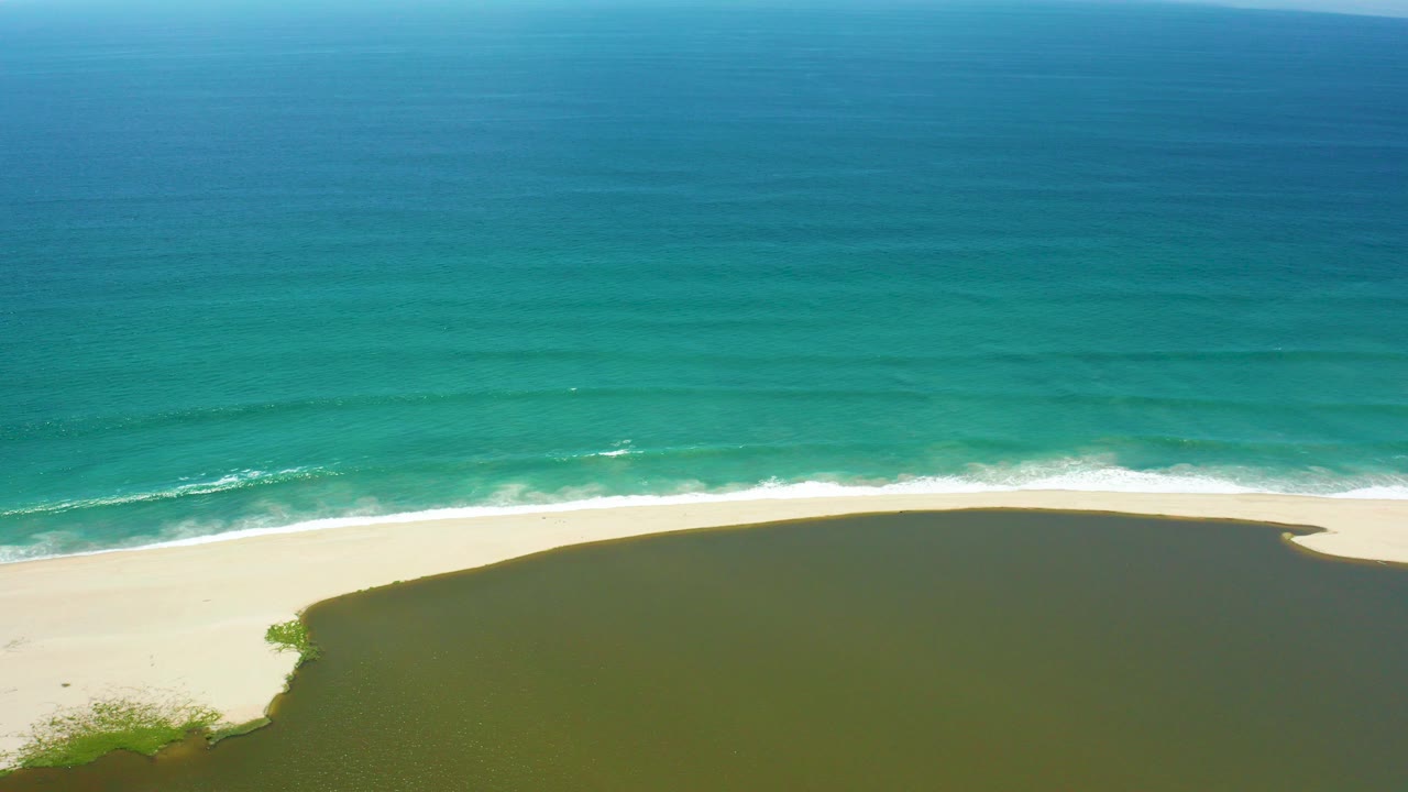 Aerial frame from San Jose del Cabo showing turquoise ocean above a thin white surf line, pale sand, and a dark green lagoon with no visible people.