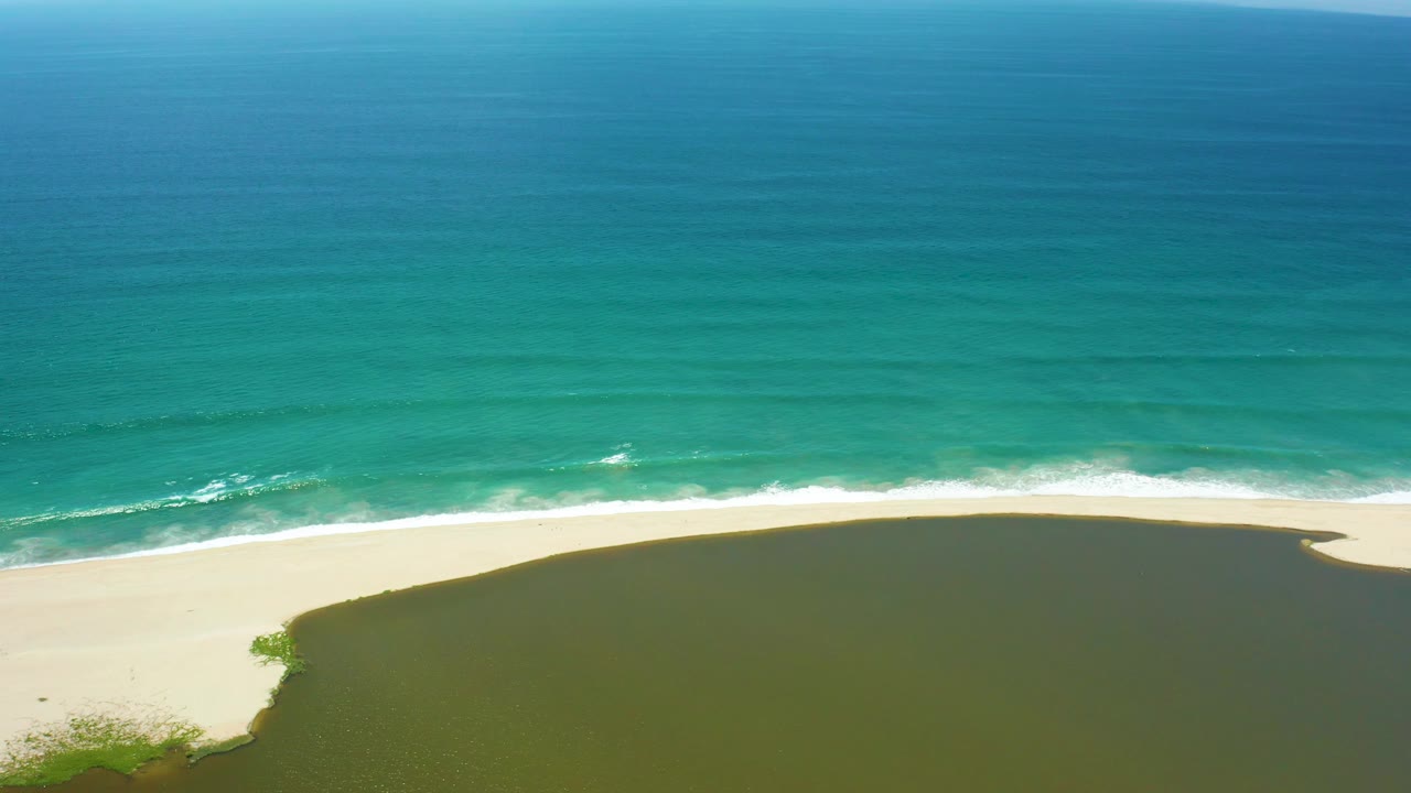 The frame holds a wide San Jose del Cabo shoreline view, with darker blue water at the top, turquoise shallows, white foam, pale sand, and lagoon water below with no visible people.