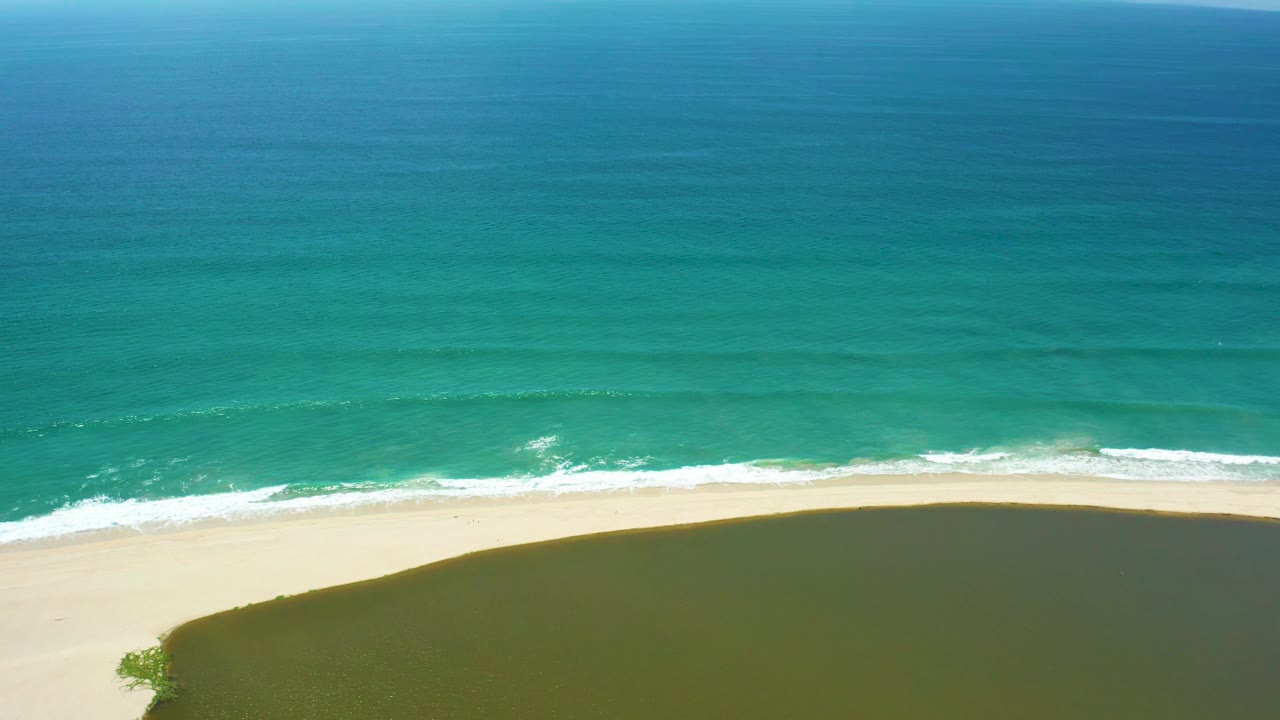 Aerial frame over San Jose Del Cabo beach with the surf band slightly lower in view, pale sand below it, darker shadow across the lower right area, and no visible people.