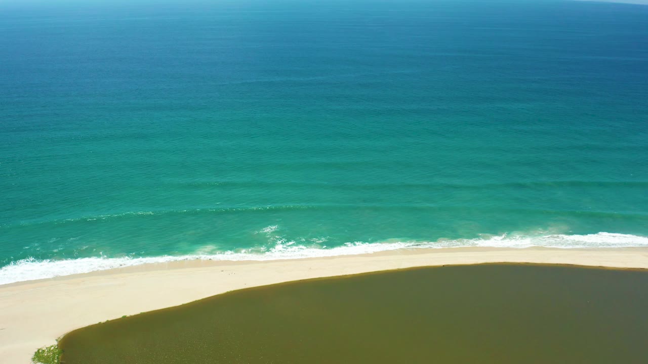 Aerial frame over San Jose Del Cabo shoreline with the white surf line running diagonally near the bottom, pale sand beneath it, dark shadow along the lower edge, and no visible people.
