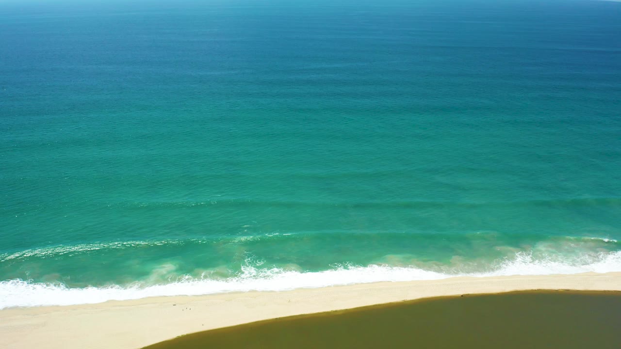 Aerial view over a broad turquoise sea with a low band of white surf breaking onto sand in San Jose del Cabo, the beach edge clear and empty of visible people.
