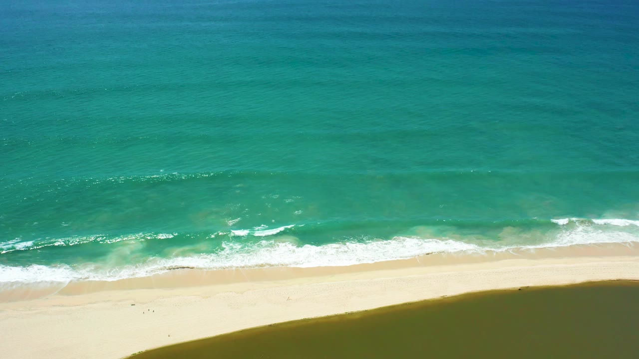Aerial San Jose Del Cabo shoreline frame with turquoise water, a low band of breaking foam, broad pale sand, and no visible people.