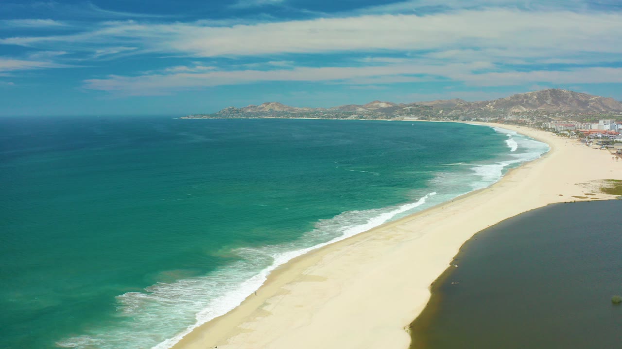 Opening frame of San Jose Del Cabo coastline with turquoise ocean on the left, pale sand curving into the distance, distant hills under clouds, and no visible people.