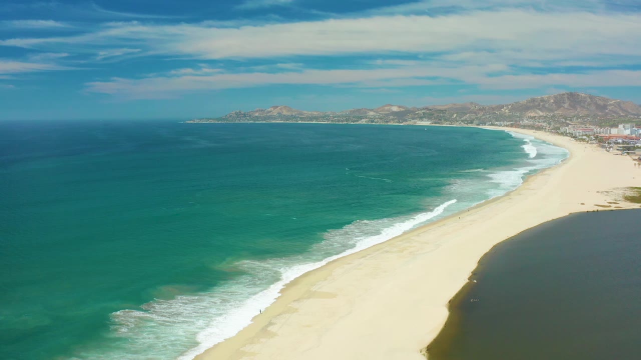 San Jose Del Cabo coastline at 0:02, with white wave bands reaching farther up the curved beach and the dark inlet edge filling the lower right, with no visible people.