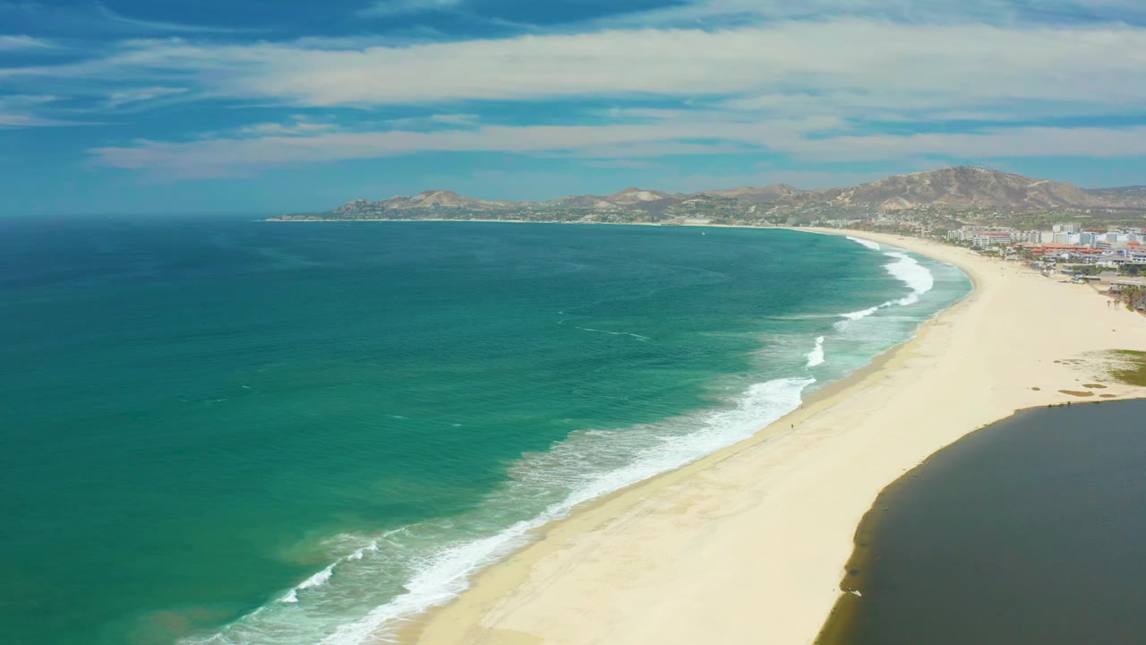 At 0:01, San Jose Del Cabo coastline remains framed from above, with small white wave lines advancing along the sand, distant hills, coastal buildings, and no visible people.