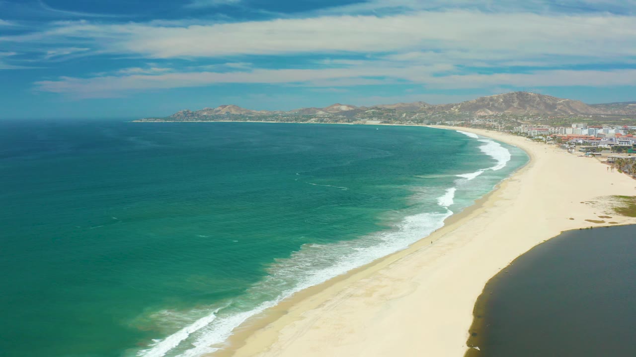 At 0:03, darker blue ocean fills more of the left frame while white surf traces the San Jose Del Cabo beach below distant hills, buildings, and no visible people.