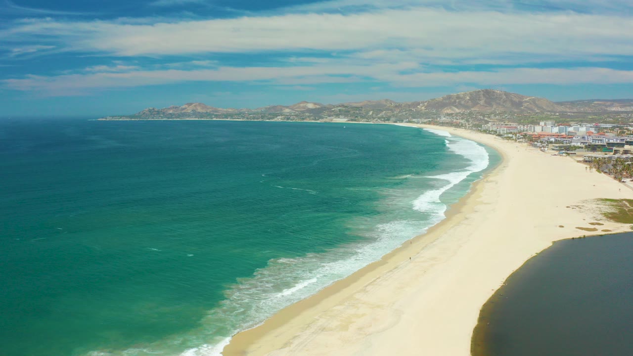 Aerial frame over San Jose Del Cabo showing deep blue water shifting to turquoise near a broad sandy beach, with distant buildings, mountains, clouds, and no visible people.