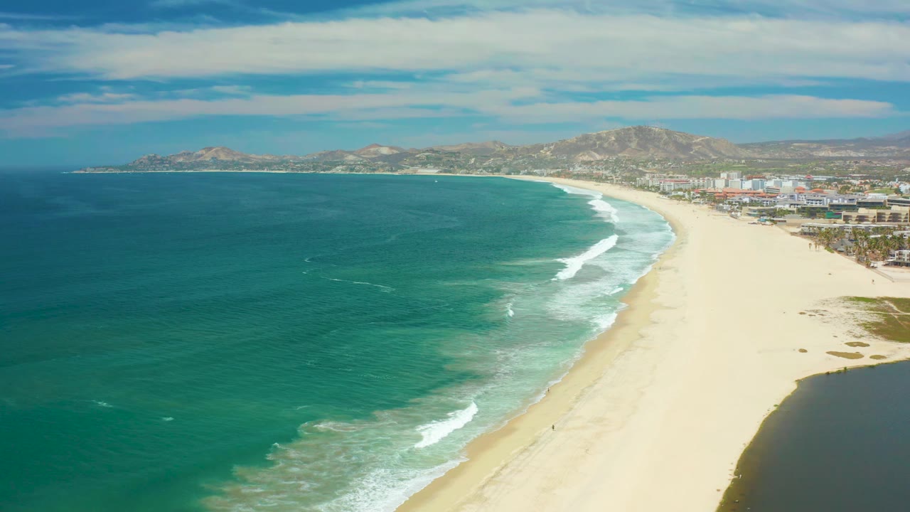 San Jose del Cabo beach seen from above as a white wave band curls along the shoreline, with teal water, pale sand, distant hills, coastal buildings, and no visible people.