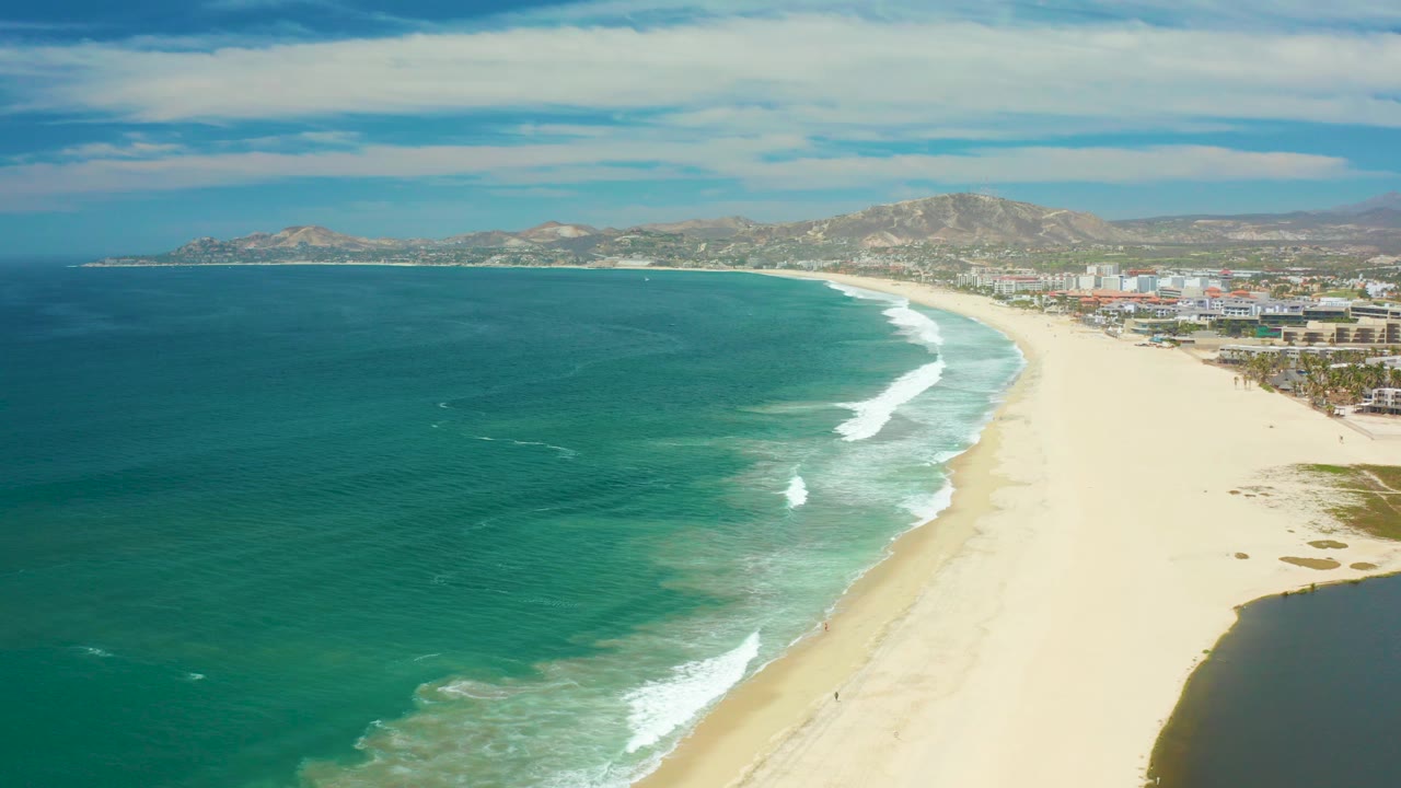 San Jose del Cabo coast with a darker teal ocean area in the foreground, fresh white surf on the sand, coastal development in the distance, hills, clouds, and no visible people.