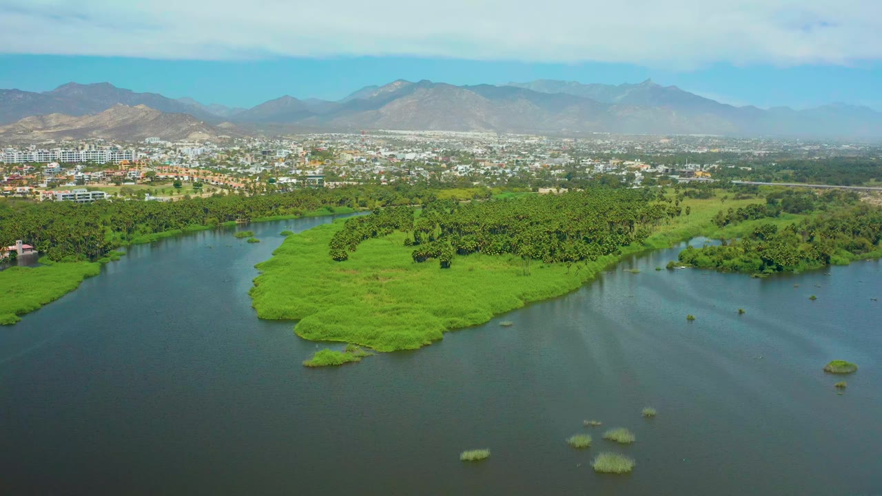 San Jose Del Cabo river landscape with a lush island centered in the water, distant urban buildings near the mountain base, and no visible people.