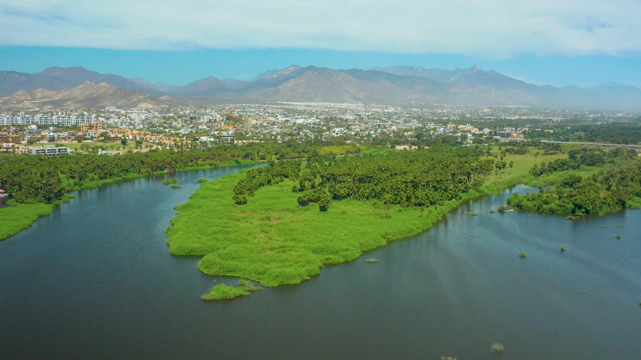 Aerial frame over San Jose del Cabo showing dark river water around bright green island vegetation, white city buildings, hazy mountains, blue sky, and no visible people.