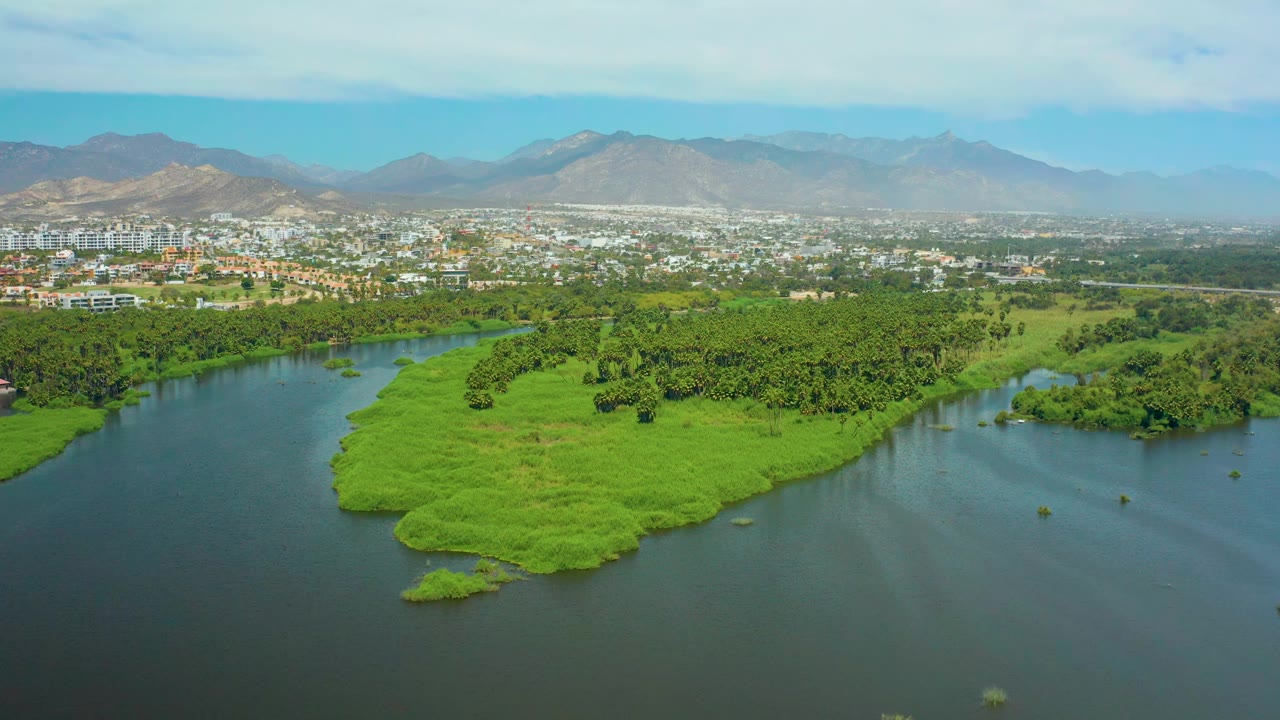 Aerial river scene with a green island, rippled water, city edge, and mountains.