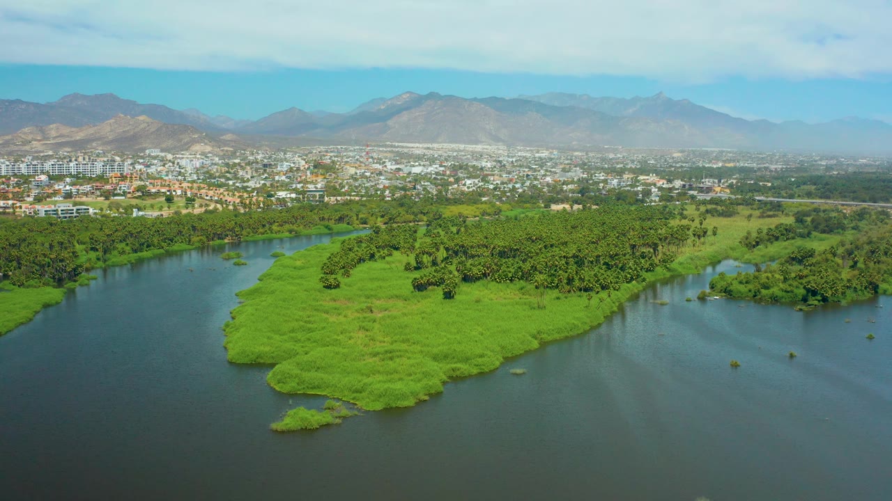 Aerial frame in San Jose del Cabo showing blue-green river water on both sides of a dense green island, low urban buildings in the distance, a hazy mountain range, and no visible people.