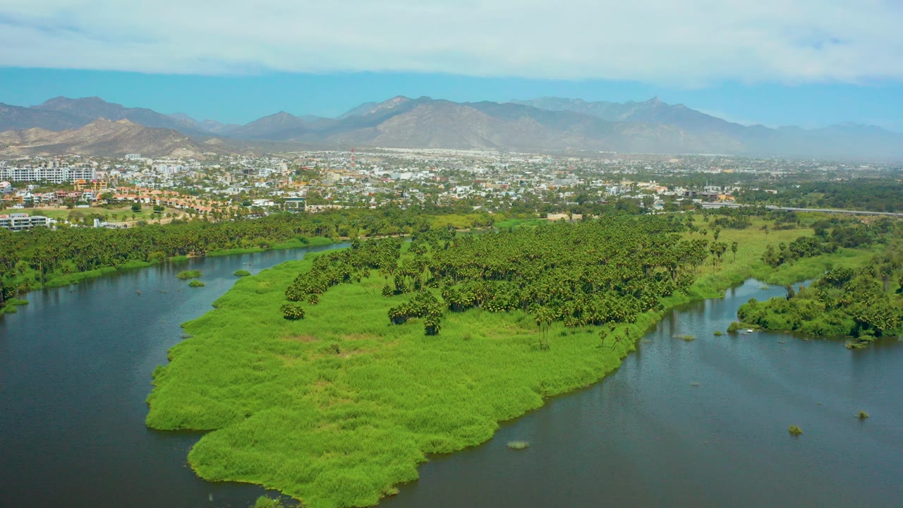 Opening aerial frame over San Jose Del Cabo, Mexico, with a bright green river island in the foreground, city buildings beyond, hazy mountains on the horizon, and no visible people.