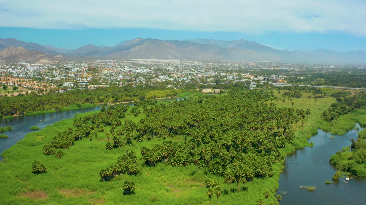 Aerial frame from San Jose del Cabo showing a winding river beside dense green fields, with a low town and hazy mountains in the distance, no visible people.