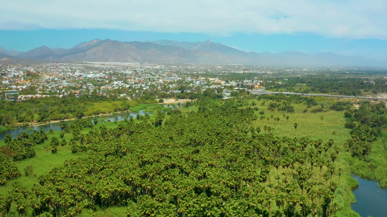 Opening aerial frame over San Jose del Cabo shows a green river corridor and fields in the foreground, a distant town, hazy mountains, blue sky, and no visible people.