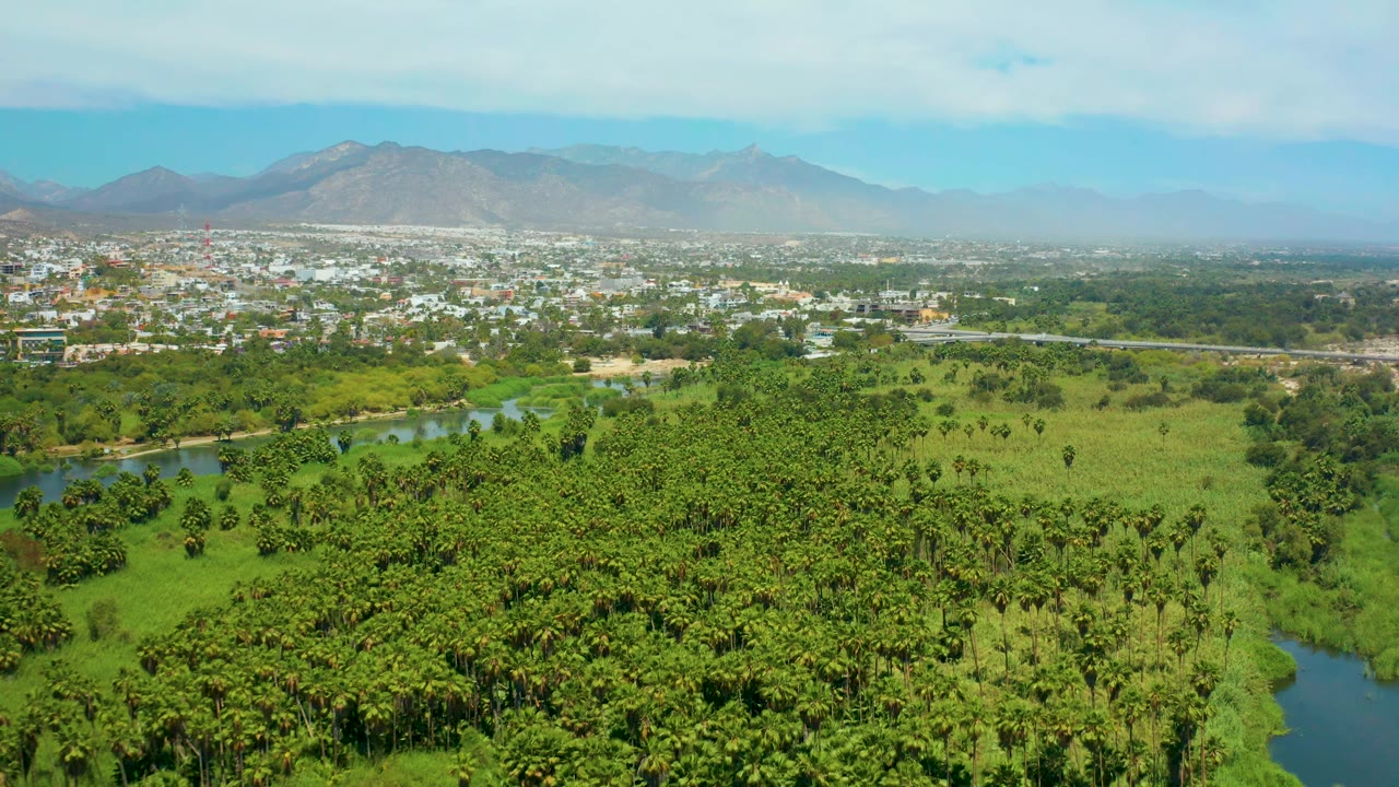 At 0:01, the San Jose del Cabo aerial view holds on river bends and dense green vegetation, with low town buildings and mountains under a pale blue sky, and no visible people.